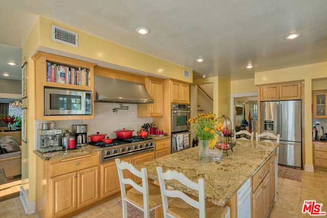 kitchen featuring a kitchen bar, light stone countertops, ventilation hood, stainless steel appliances, and a kitchen island with sink