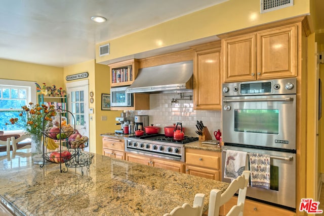 kitchen featuring wall chimney exhaust hood, light stone countertops, backsplash, and appliances with stainless steel finishes