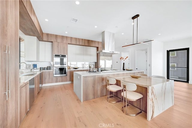 kitchen featuring white cabinetry, island range hood, a kitchen island with sink, hanging light fixtures, and sink