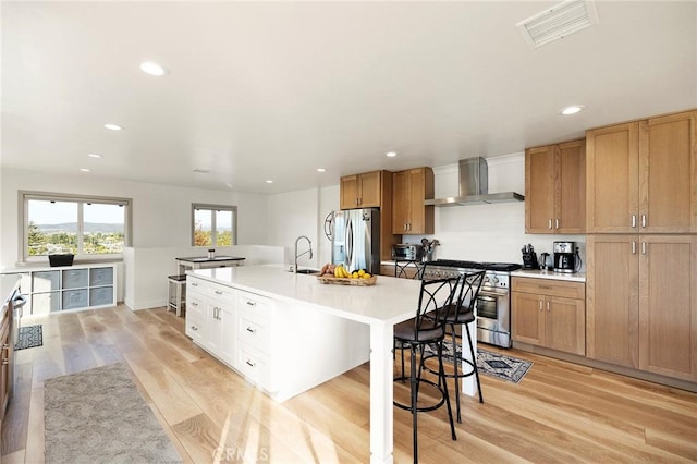 kitchen with sink, wall chimney exhaust hood, light wood-type flooring, an island with sink, and stainless steel appliances