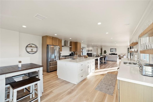 kitchen featuring white cabinets, wall chimney exhaust hood, stainless steel fridge, light wood-type flooring, and an island with sink