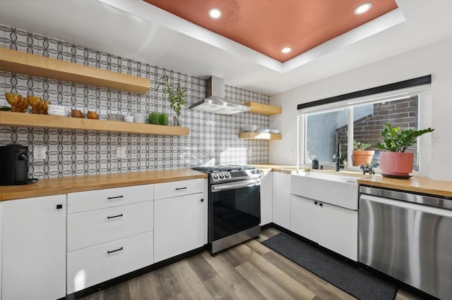 kitchen featuring appliances with stainless steel finishes, backsplash, wall chimney range hood, butcher block countertops, and white cabinetry