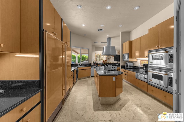 kitchen with sink, built in appliances, dark stone countertops, island exhaust hood, and an island with sink