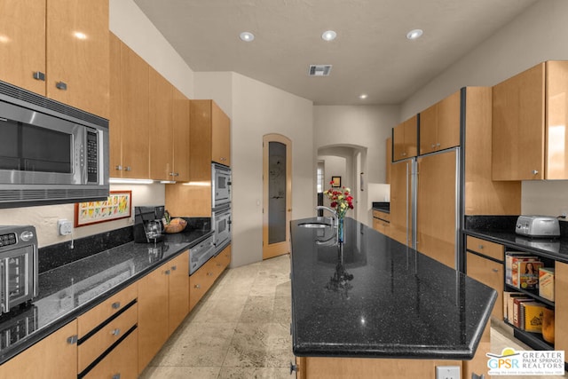 kitchen with built in appliances, sink, a center island with sink, and dark stone countertops