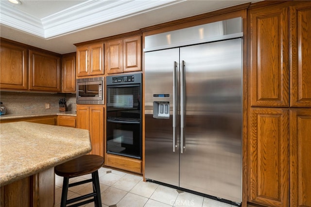 kitchen featuring a kitchen breakfast bar, built in appliances, decorative backsplash, light tile patterned floors, and ornamental molding