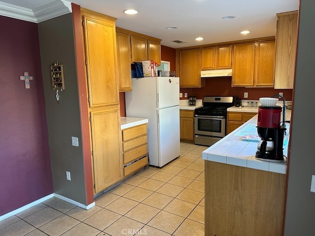 kitchen featuring stainless steel gas range oven, white refrigerator, tile countertops, crown molding, and light tile patterned flooring