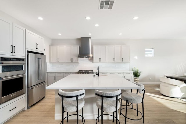 kitchen featuring white cabinets, wall chimney exhaust hood, an island with sink, and stainless steel appliances