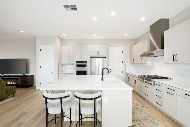 kitchen with a center island with sink, sink, white cabinetry, appliances with stainless steel finishes, and wall chimney exhaust hood