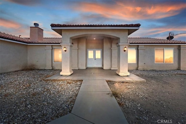 exterior entry at dusk featuring french doors, stucco siding, a chimney, and a tile roof