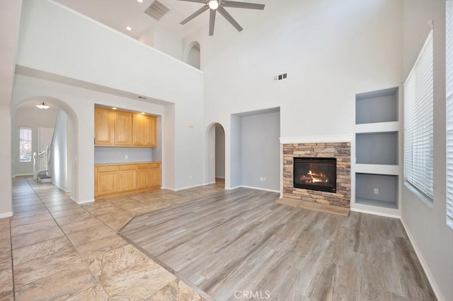 unfurnished living room featuring a stone fireplace, light hardwood / wood-style flooring, built in shelves, ceiling fan, and a towering ceiling