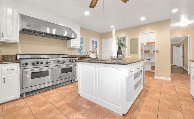 kitchen with wall chimney exhaust hood, ceiling fan, sink, range with two ovens, and a center island