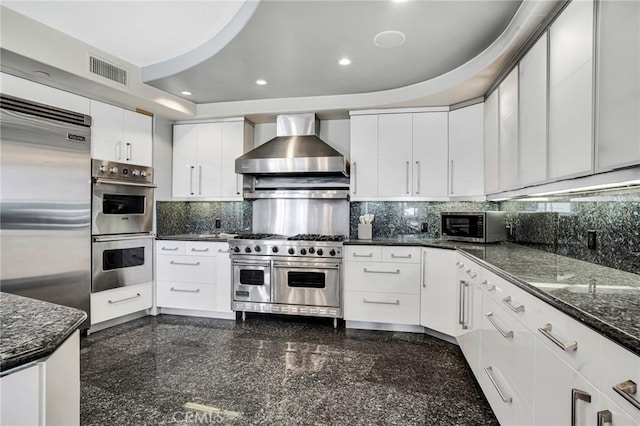 kitchen featuring wall chimney range hood, backsplash, dark stone counters, white cabinets, and high end appliances