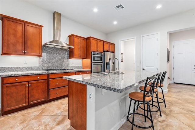 kitchen featuring sink, wall chimney exhaust hood, stainless steel appliances, light stone counters, and an island with sink