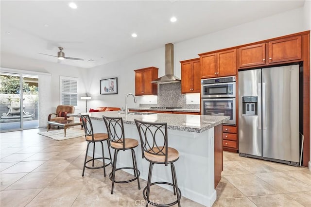 kitchen with a center island with sink, appliances with stainless steel finishes, wall chimney range hood, a kitchen bar, and a sink