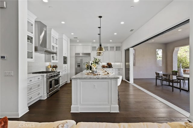 kitchen featuring a kitchen island, wall chimney range hood, built in appliances, white cabinetry, and hanging light fixtures