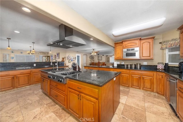 kitchen featuring appliances with stainless steel finishes, wall chimney exhaust hood, ceiling fan, a kitchen island with sink, and hanging light fixtures