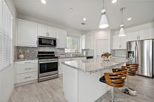 kitchen with appliances with stainless steel finishes, a center island, light stone counters, white cabinets, and decorative light fixtures