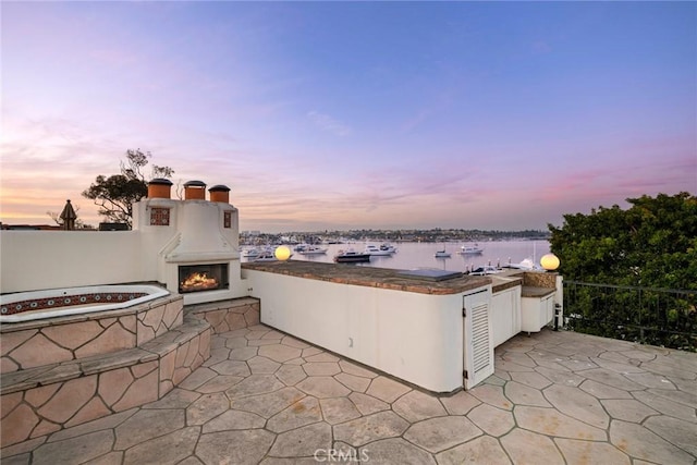 patio terrace at dusk with area for grilling, a water view, and exterior fireplace