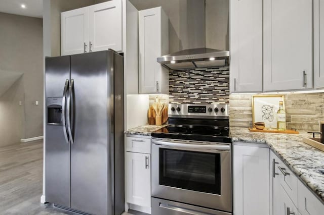 kitchen with white cabinetry, backsplash, light stone counters, stainless steel appliances, and wall chimney range hood