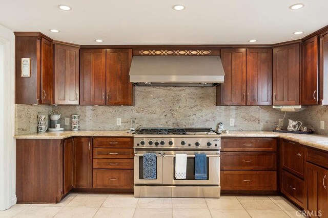 kitchen with light stone countertops, wall chimney exhaust hood, range with two ovens, tasteful backsplash, and light tile patterned flooring