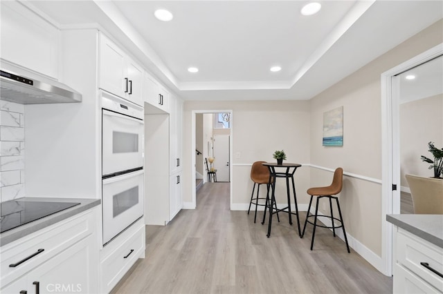 kitchen featuring light wood-type flooring, black electric cooktop, double oven, a tray ceiling, and white cabinetry