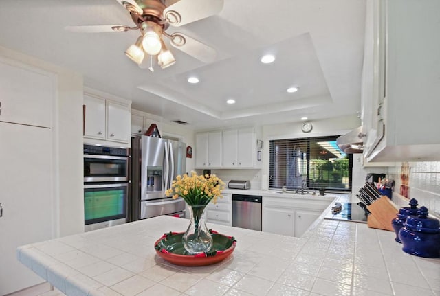 kitchen featuring a raised ceiling, tile countertops, kitchen peninsula, white cabinets, and appliances with stainless steel finishes