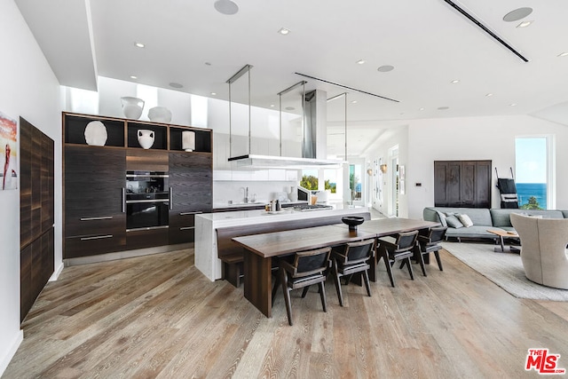 kitchen with a breakfast bar, a center island with sink, wall chimney range hood, decorative light fixtures, and light hardwood / wood-style floors