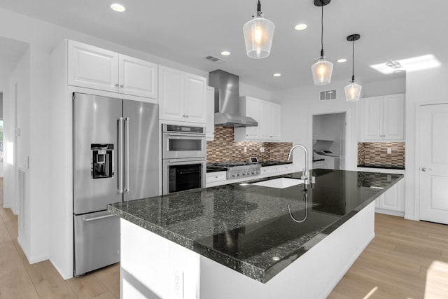 kitchen featuring appliances with stainless steel finishes, a kitchen island with sink, wall chimney range hood, light hardwood / wood-style floors, and white cabinetry