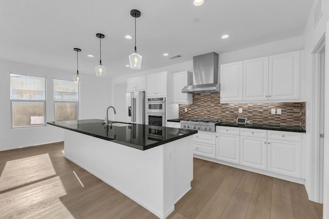 kitchen with pendant lighting, a center island with sink, wall chimney exhaust hood, white cabinetry, and stainless steel appliances