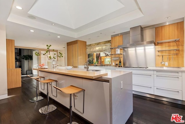 kitchen featuring a raised ceiling, a center island with sink, white cabinetry, and wall chimney range hood