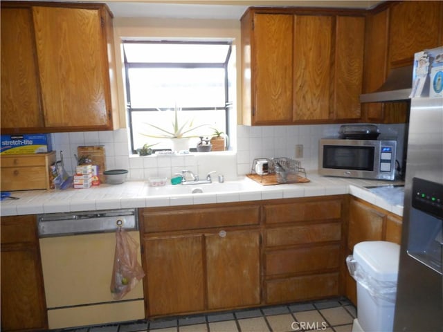 kitchen featuring tile countertops, backsplash, exhaust hood, sink, and appliances with stainless steel finishes