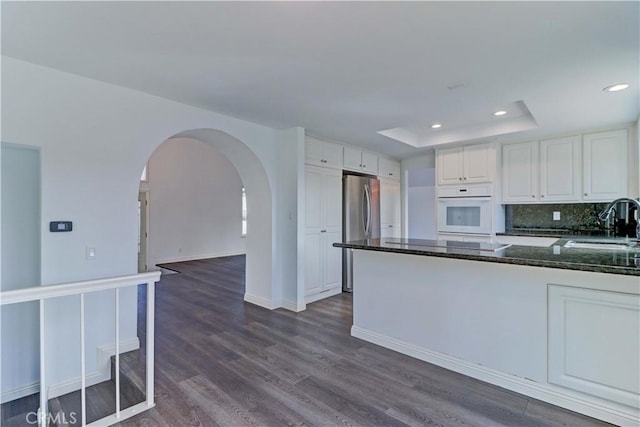 kitchen with white oven, white cabinetry, a tray ceiling, dark stone counters, and sink