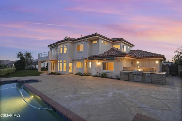 back house at dusk featuring an outdoor kitchen, pool water feature, a patio area, and a balcony