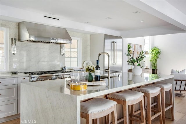 kitchen with light wood-type flooring, an island with sink, high end fridge, and wall chimney range hood