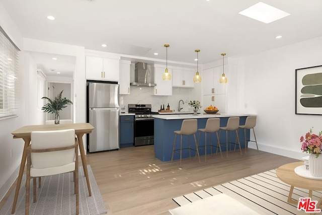 kitchen with wall chimney exhaust hood, stainless steel appliances, light hardwood / wood-style flooring, white cabinets, and hanging light fixtures