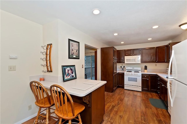 kitchen with dark brown cabinetry, dark hardwood / wood-style floors, kitchen peninsula, white appliances, and a breakfast bar area