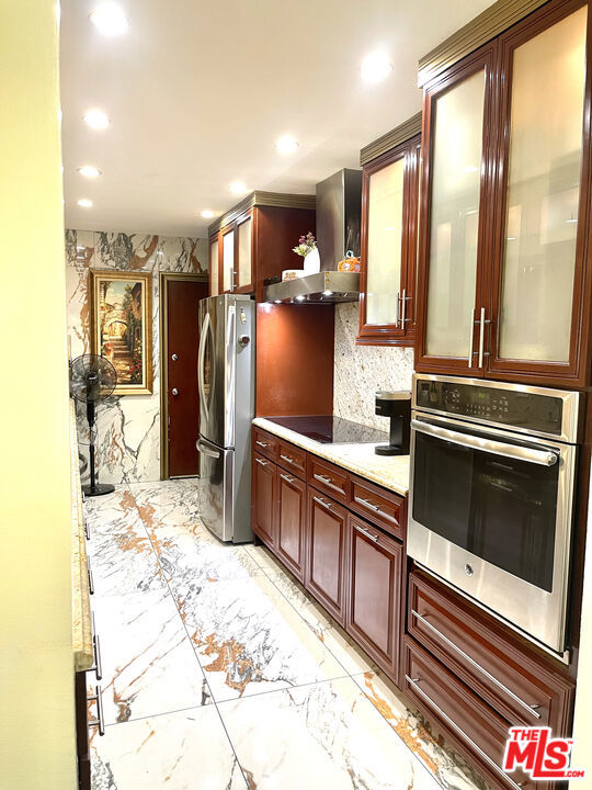 kitchen with decorative backsplash, stainless steel appliances, light stone counters, and wall chimney range hood