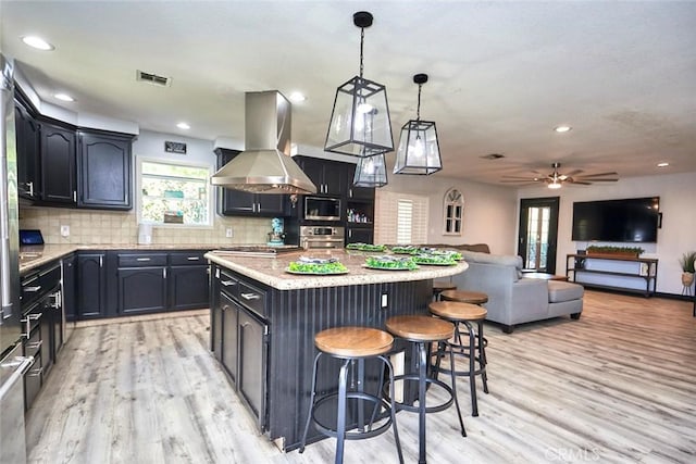 kitchen with wall chimney range hood, a kitchen island, appliances with stainless steel finishes, decorative light fixtures, and light hardwood / wood-style floors