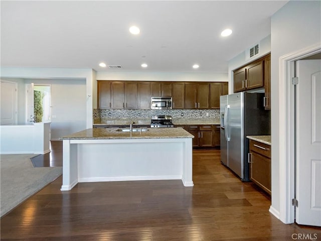 kitchen featuring dark wood-type flooring, decorative backsplash, light stone countertops, an island with sink, and stainless steel appliances