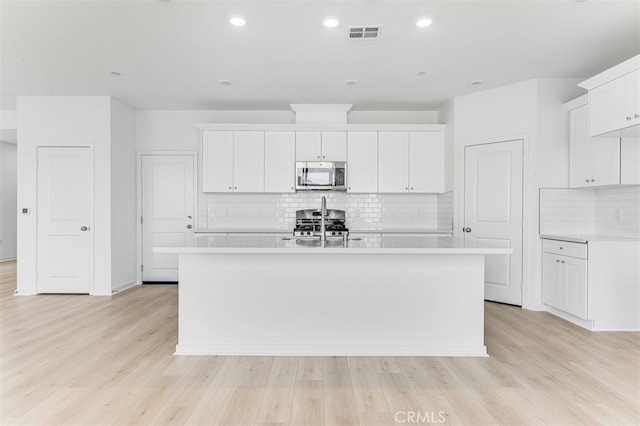 kitchen featuring light countertops, stainless steel microwave, an island with sink, and white cabinetry