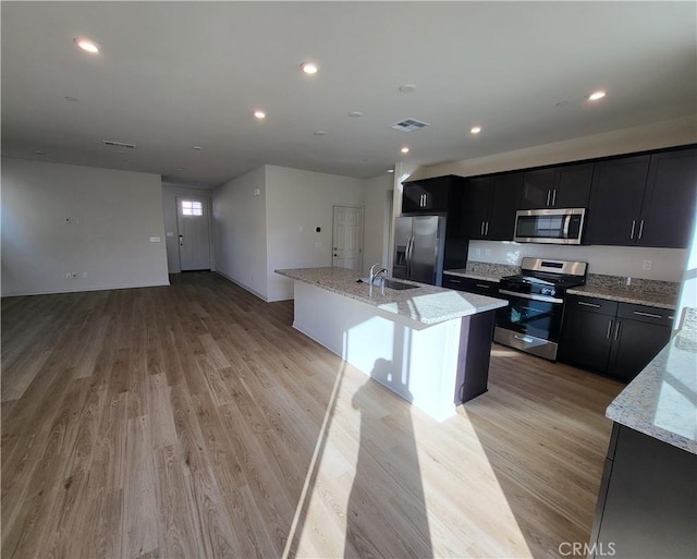 kitchen featuring light stone countertops, light wood-type flooring, stainless steel appliances, a kitchen island with sink, and sink