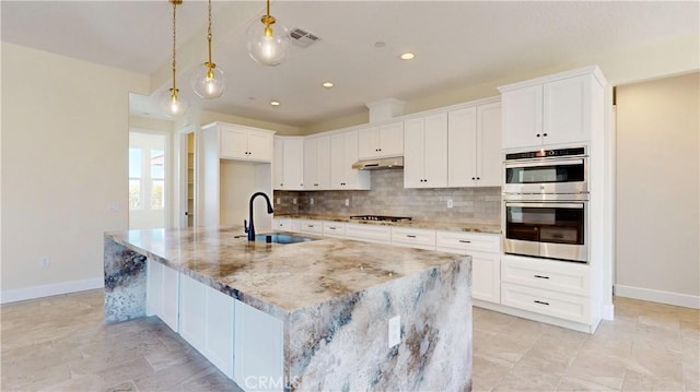 kitchen with appliances with stainless steel finishes, a kitchen island with sink, sink, white cabinets, and hanging light fixtures