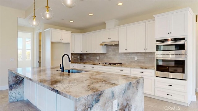 kitchen with light stone counters, a center island with sink, decorative light fixtures, and sink