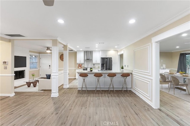 kitchen featuring stainless steel refrigerator with ice dispenser, wall chimney exhaust hood, light wood-type flooring, white cabinetry, and a breakfast bar area