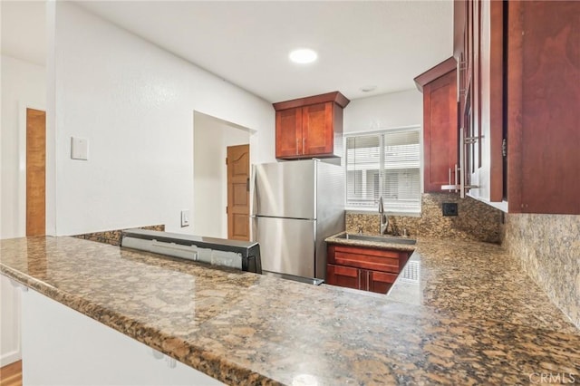 kitchen featuring backsplash, dark stone countertops, kitchen peninsula, sink, and stainless steel refrigerator