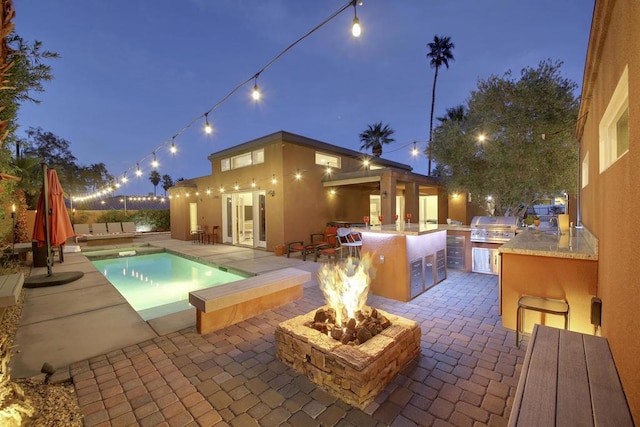 pool at twilight with area for grilling, a fire pit, grilling area, and a patio area