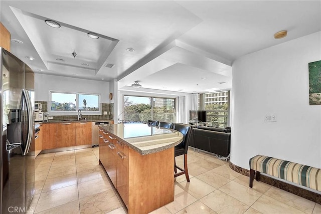 kitchen featuring a center island, a raised ceiling, stainless steel dishwasher, refrigerator with ice dispenser, and a breakfast bar area