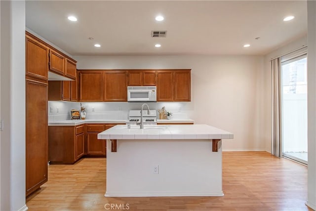 kitchen with white microwave, an island with sink, tile counters, and brown cabinets