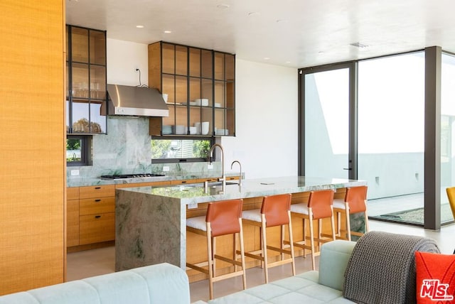 kitchen with a breakfast bar, a center island with sink, wall chimney range hood, sink, and tasteful backsplash