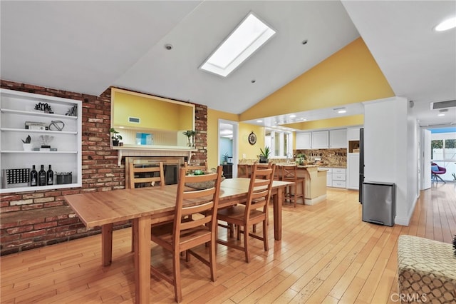 dining area featuring a brick fireplace, a skylight, light hardwood / wood-style floors, and high vaulted ceiling